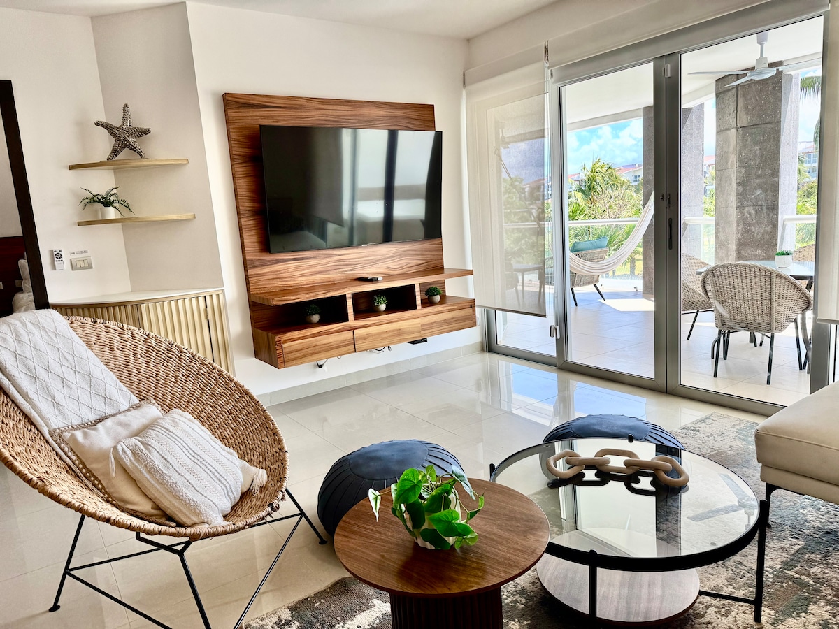A cozy living area features a large wall-mounted television and stylish wooden cabinetry. Comfortable seating options include a woven chair and a round coffee table adorned with a small plant. Natural light streams in through glass doors leading to a balcony, enhancing the space.