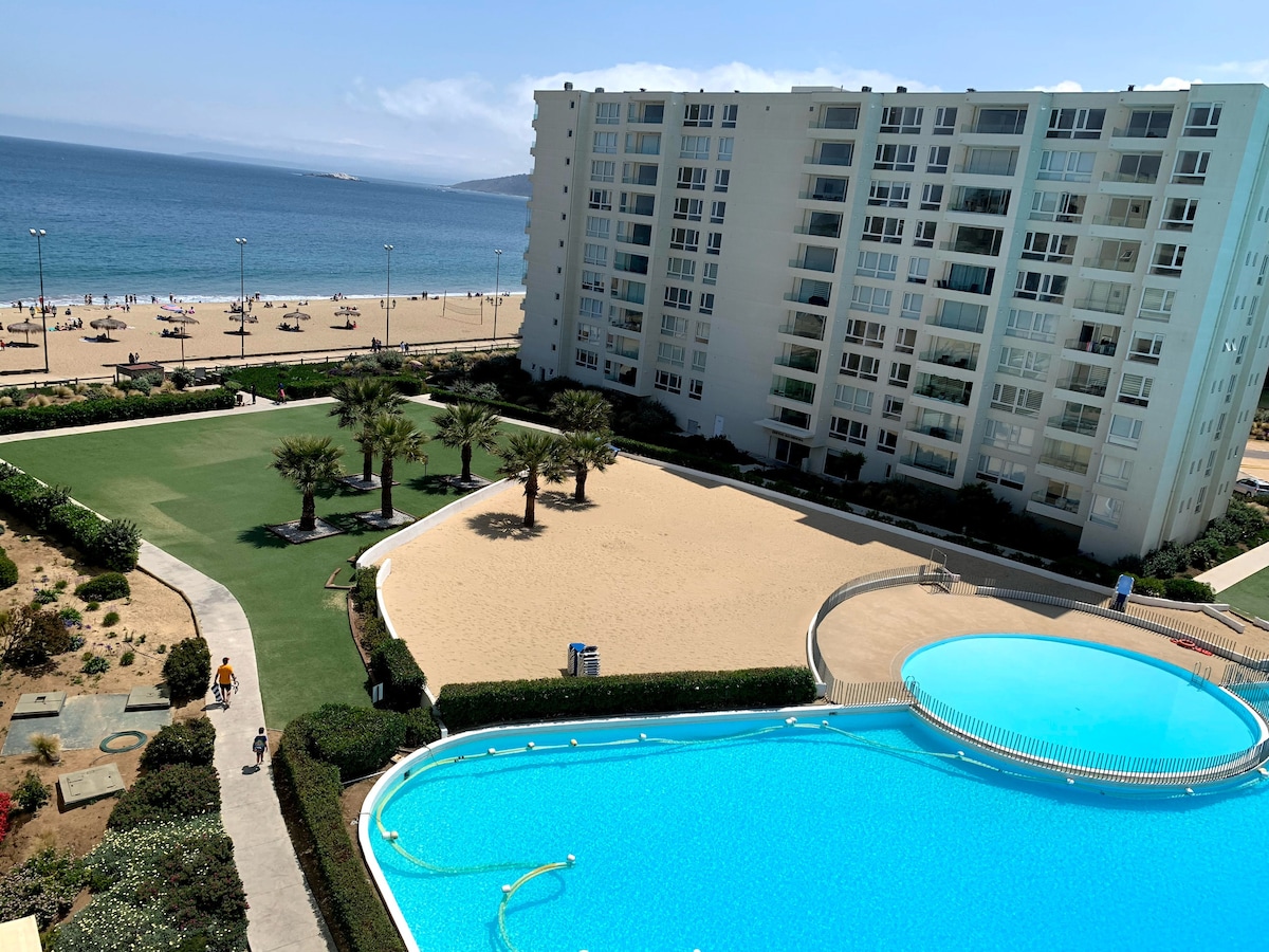 The image captures a panoramic view of a large pool surrounded by landscaped gardens and sandy areas. Beyond, the ocean is visible, enhancing the serene atmosphere. Sun loungers are arranged along the beach, inviting relaxation under clear skies.