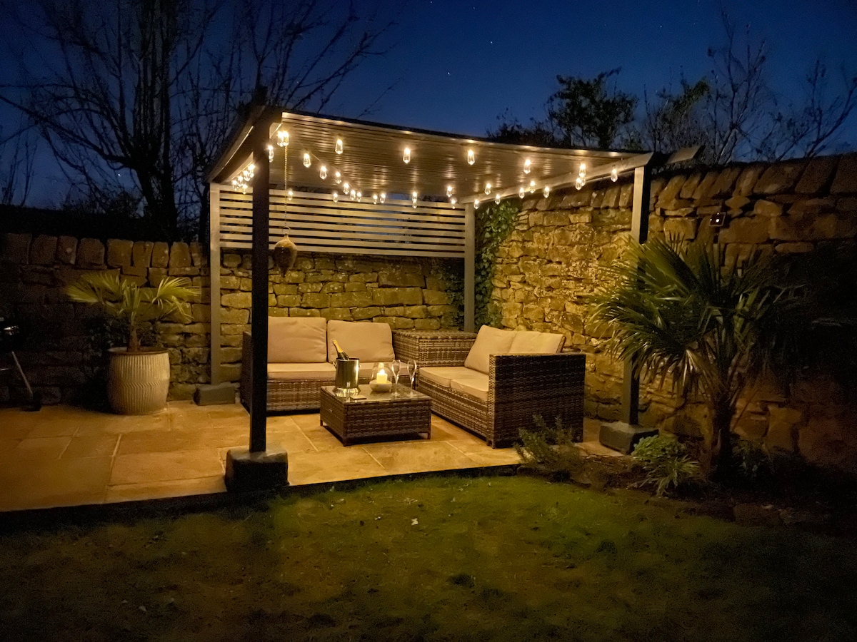 An outdoor seating area is illuminated by string lights, creating a serene atmosphere at night. The space features comfortable wicker seating and a coffee table, surrounded by natural stone walls and a landscaped garden. Potted plants add a touch of greenery to the setting.