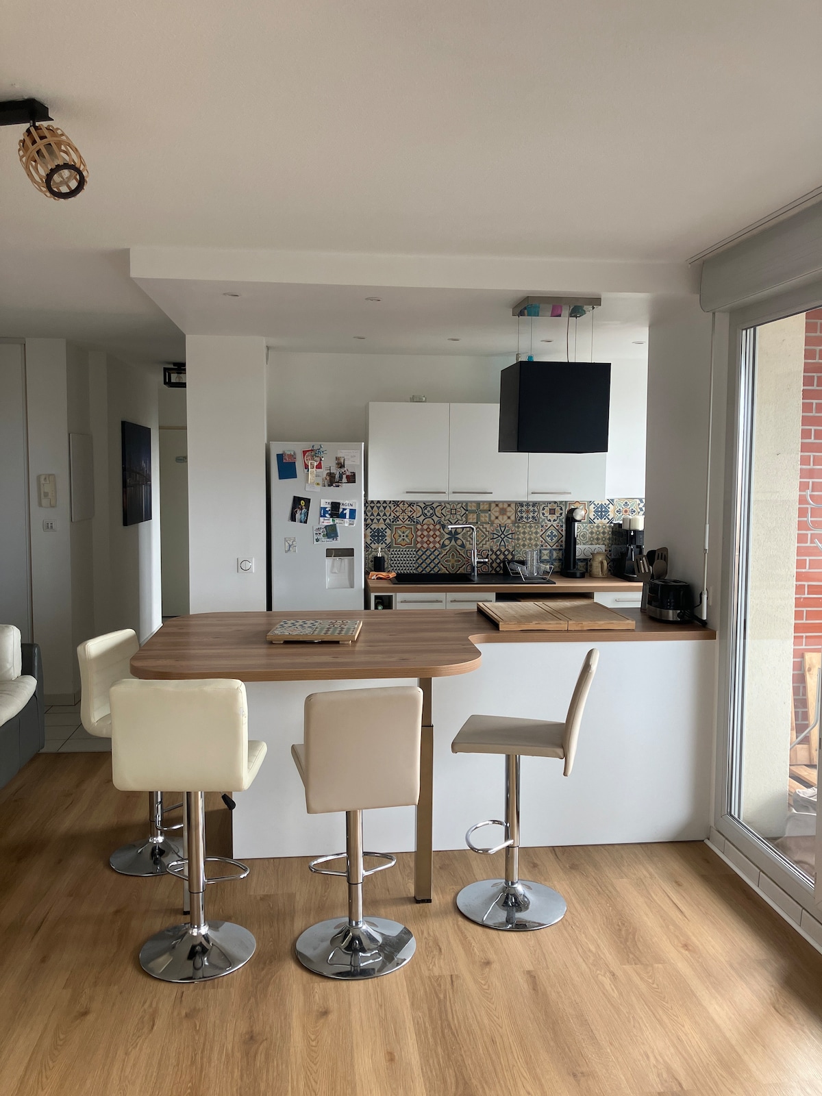 A modern kitchen features a stylish breakfast bar with four high-backed stools. Decorative tiles adorn the backsplash, complementing the wooden countertop. Natural light floods the space through large windows, enhancing the overall bright and open atmosphere.