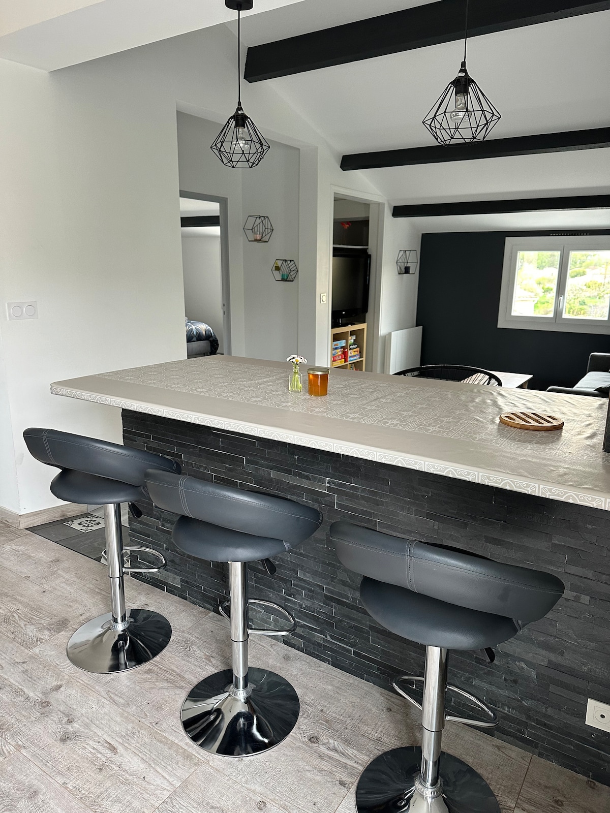 A kitchen counter features three sleek black bar stools with chrome bases. Modern pendant lights hang above, providing ambient lighting. A small vase with a flower rests on the counter, alongside a drink. The background reveals a cozy living area with large windows.