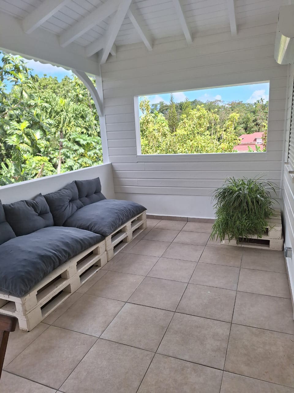 A covered terrace is furnished with a comfortable seating area made from wooden pallets, adorned with plush gray cushions. A small potted plant adds a touch of greenery, while large windows offer views of the surrounding trees and nearby rooftops.