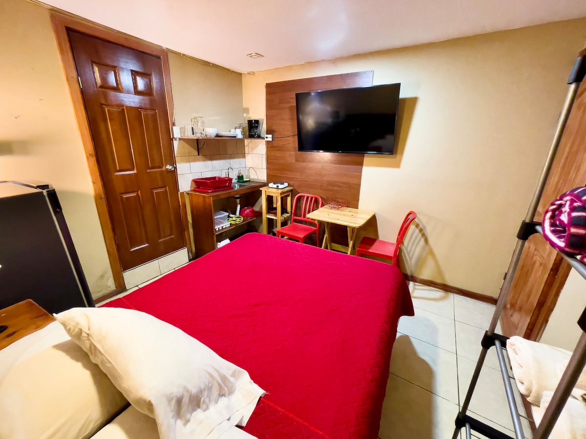 A cozy room features a spacious bed covered in a red quilt, complemented by neutral-colored walls. A flat-screen television is mounted on the wall across from the bed. A small dining area with two chairs and a table, along with shelves for storage, is also present.