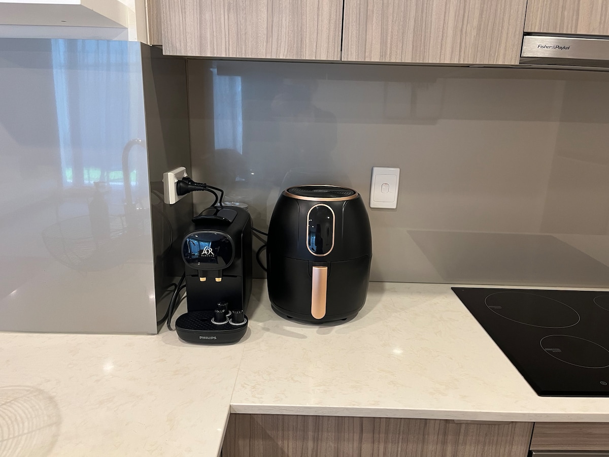 A modern kitchen countertop displays a coffee machine and an air fryer, both featuring sleek black designs with gold accents. The surface is light-colored, complementing the cabinetry. A smooth cooktop extends along the countertop, noting a tidy and functional preparation area.
