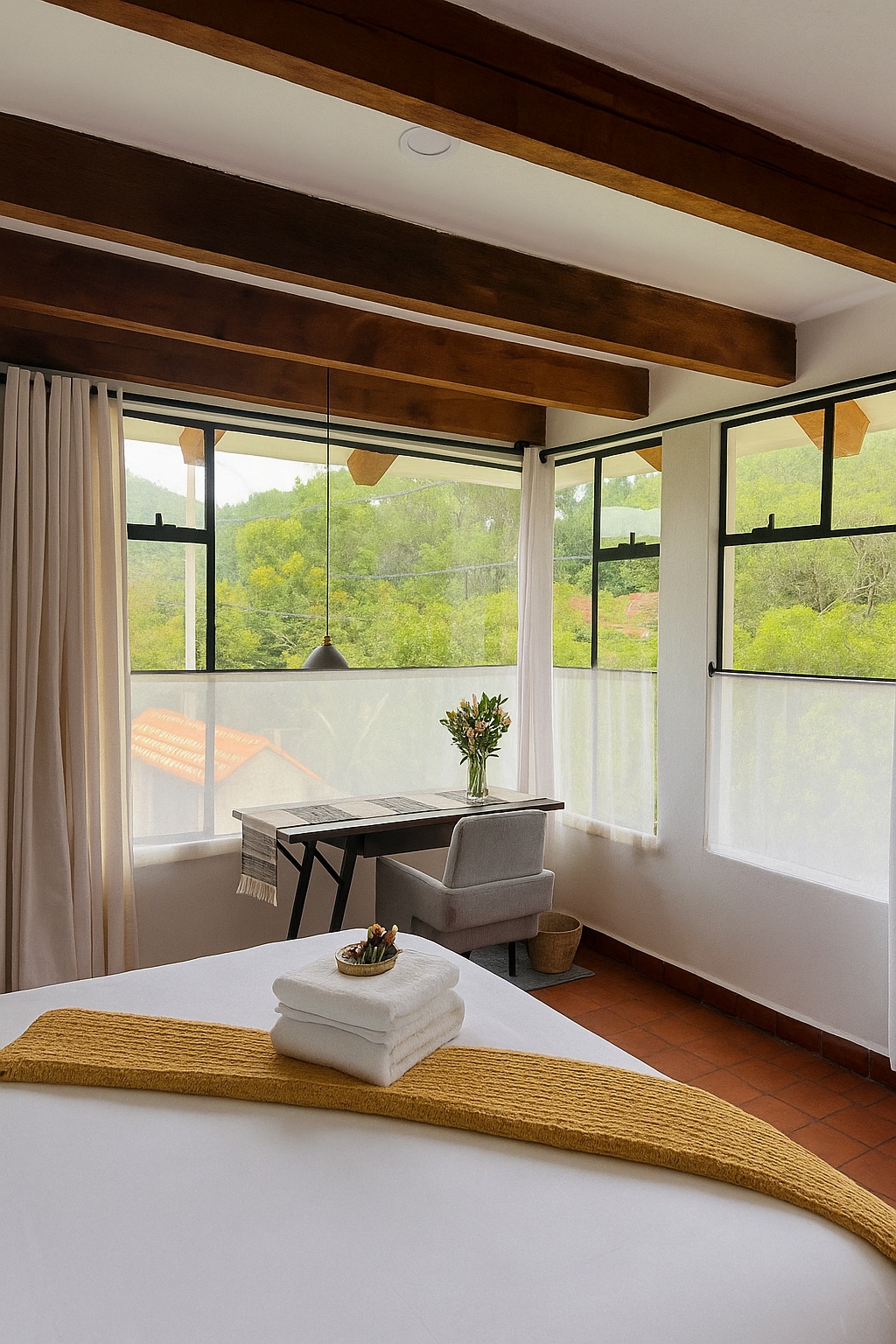 A bright sleeping area features large windows that overlook lush greenery. Wooden beams span the ceiling, adding warmth to the space. A simple desk and chair are positioned by the window, complemented by a small vase of fresh flowers placed on the desk.