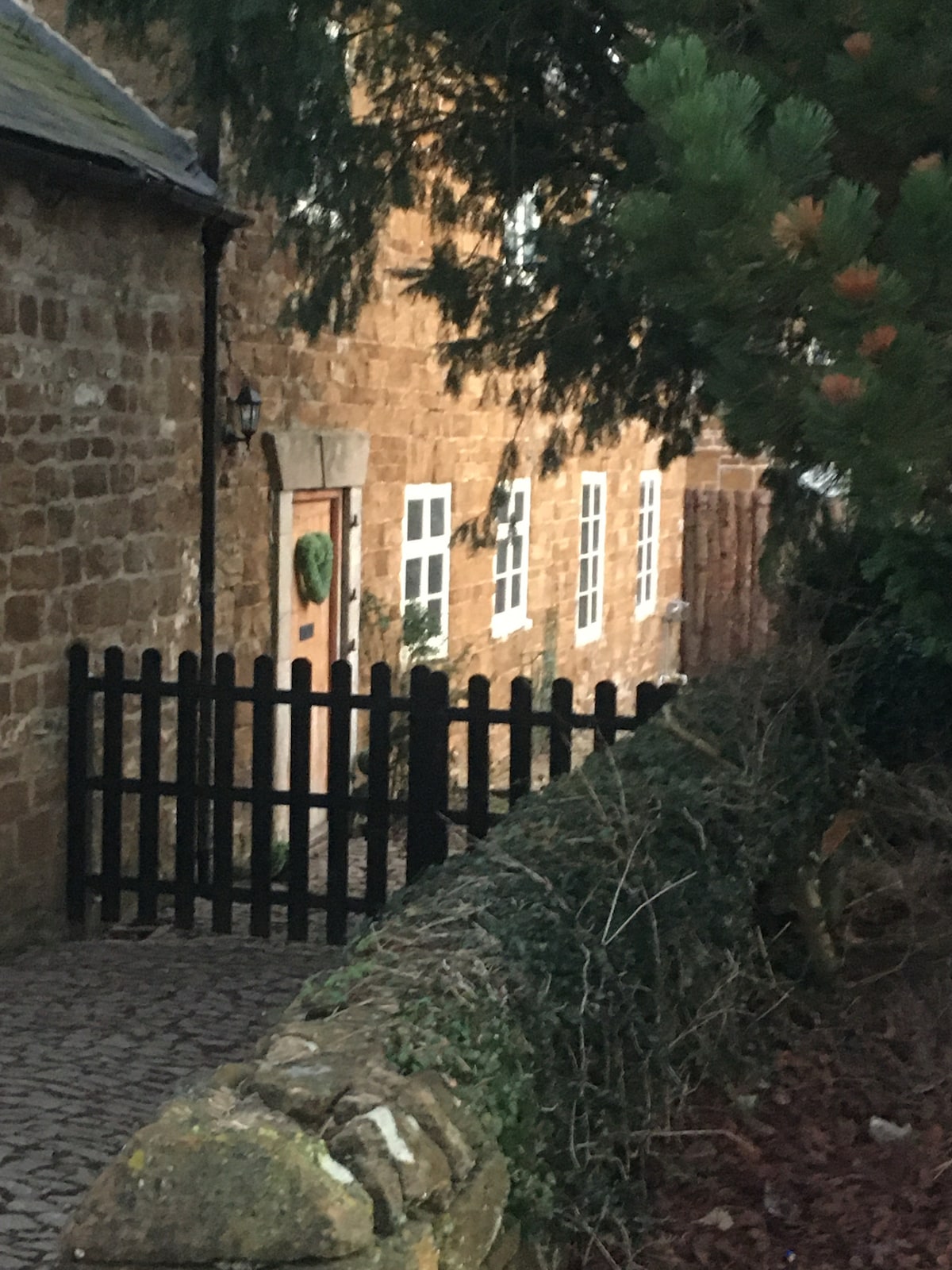 A charming stone cottage is portrayed, framed by a wooden gate and a low stone wall. Several white windows reflect soft light against the warm stone facade. Lush greenery partially obscures the view, adding to the inviting atmosphere of the setting.