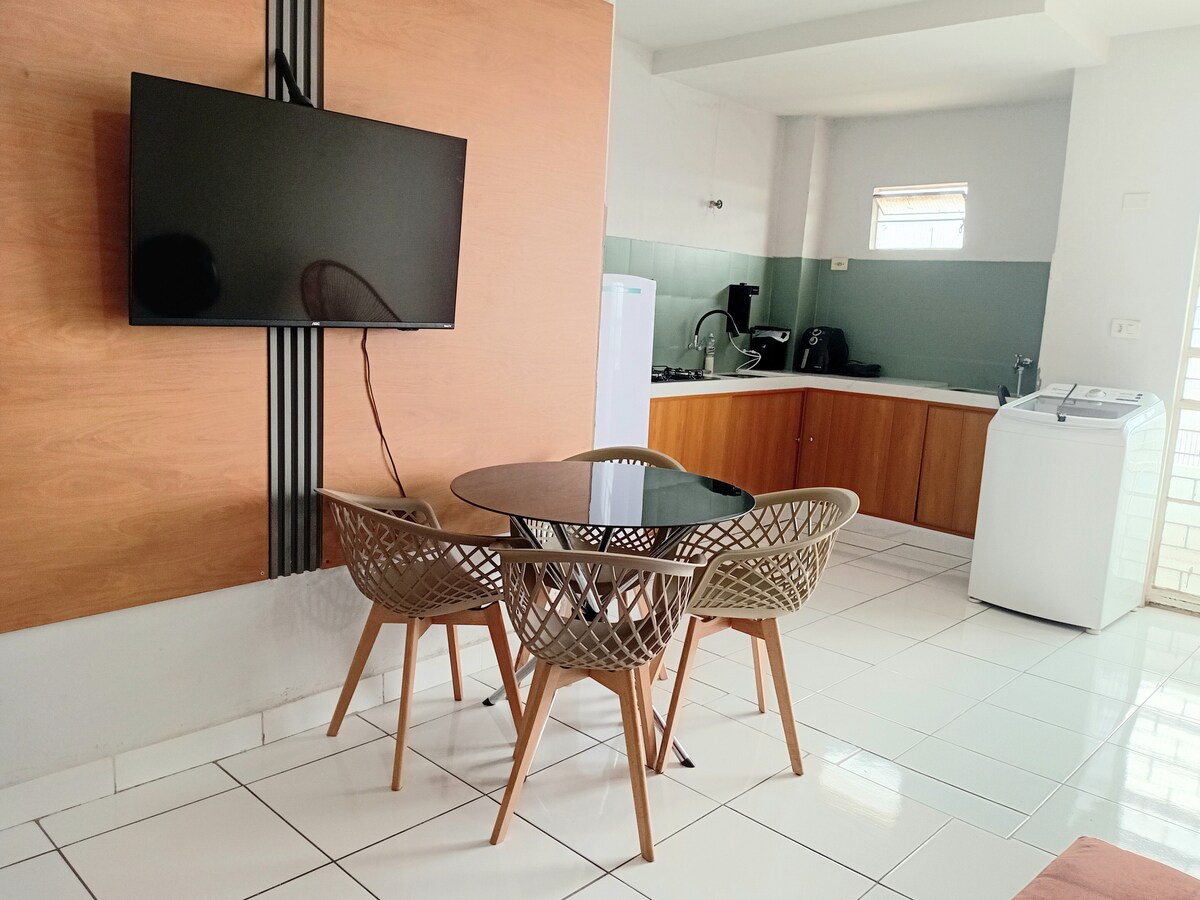A dining area features a round black table surrounded by four beige woven chairs, set against a modern wood-paneled wall. A flat-screen TV is mounted nearby, while a functional kitchen area with a refrigerator and sink is visible in the background, complemented by white tiled flooring.