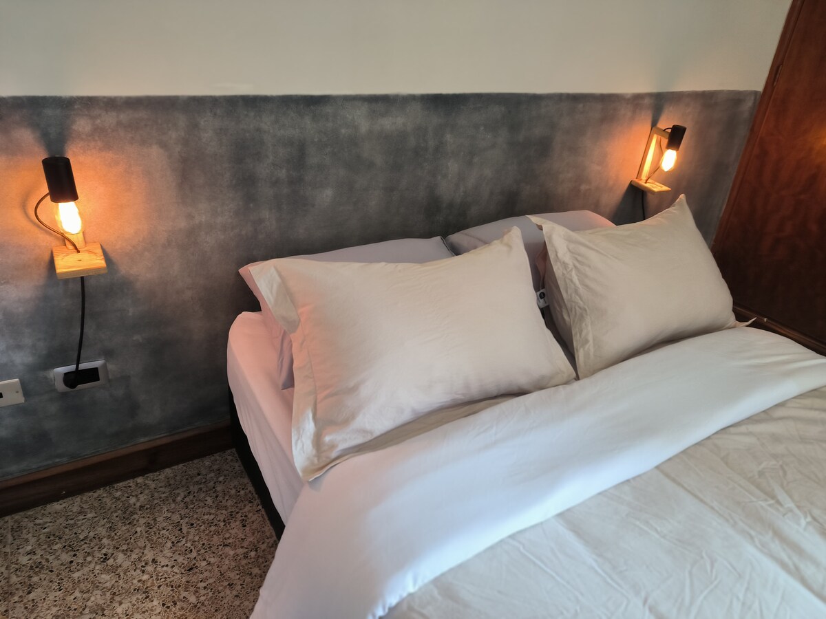 A spacious bed is positioned against a textured gray wall, complemented by warm lighting from two wall-mounted lamps on either side. Soft, light-colored bedding enhances the calming ambiance of the room.
