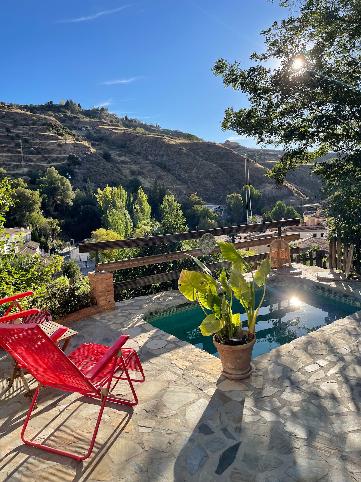 A private terrace features a small pool surrounded by natural stone flooring and vibrant greenery. Red lounge chairs are positioned to enjoy views of the surrounding hills. Sunlight creates a warm glow, enhancing the tranquil atmosphere of the space.