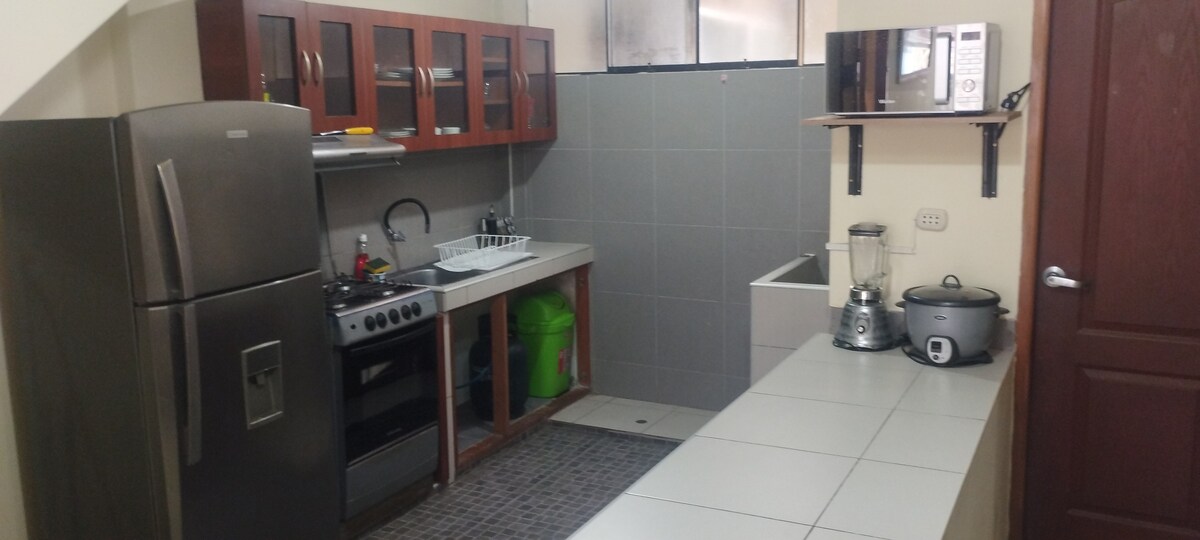 A functional kitchen is equipped with dark wooden cabinets and stainless steel appliances. A refrigerator, stove, and sink are arranged along the counter. A blender and rice cooker are visible, with a spacious counter surface providing ample workspace.