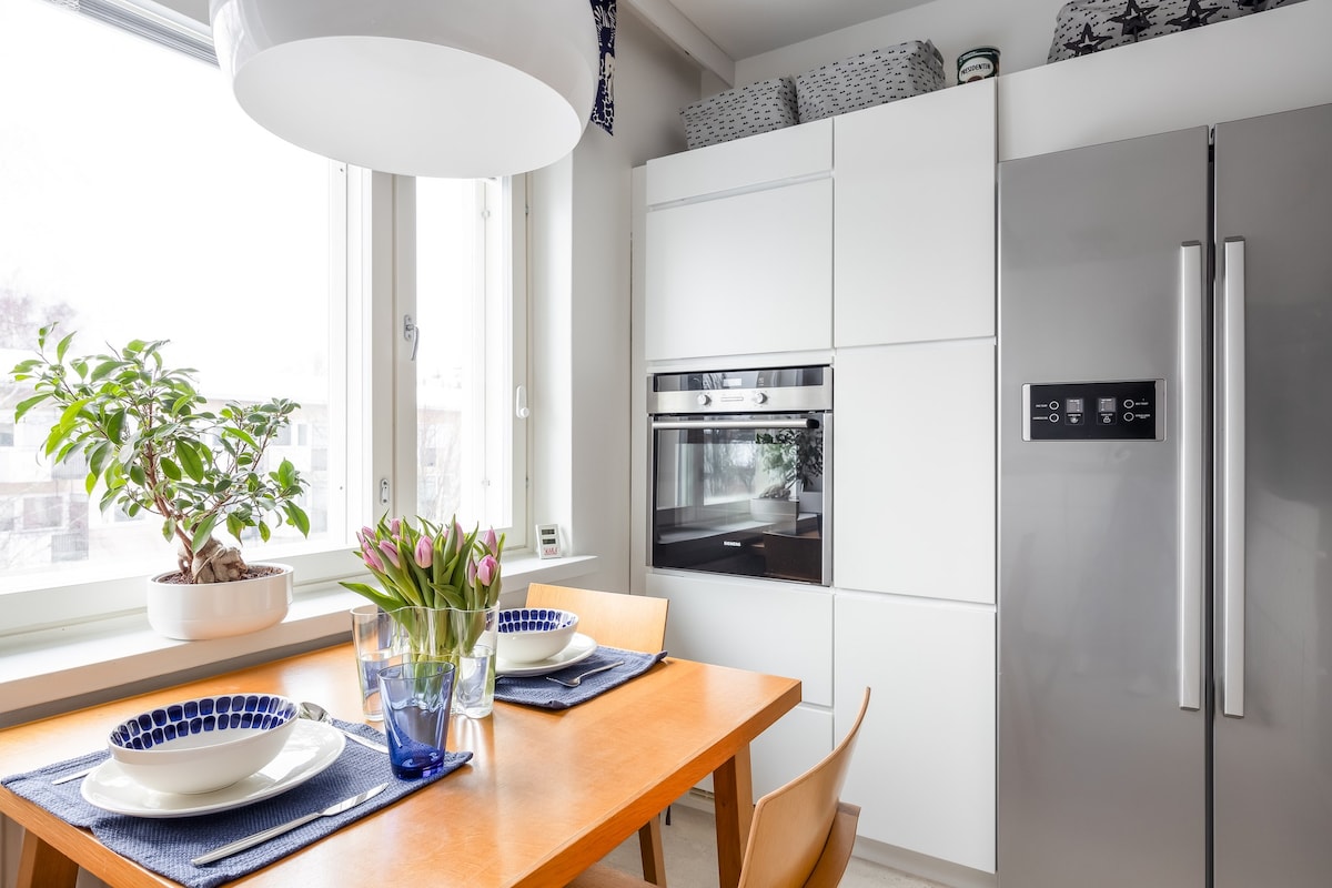 A modern kitchen area is highlighted with a wooden dining table set for two, featuring blue and white dishes. Fresh tulips in a clear vase add a touch of color. Sleek white cabinetry and a stainless steel refrigerator complete the clean, functional design, with natural light streaming through large windows.