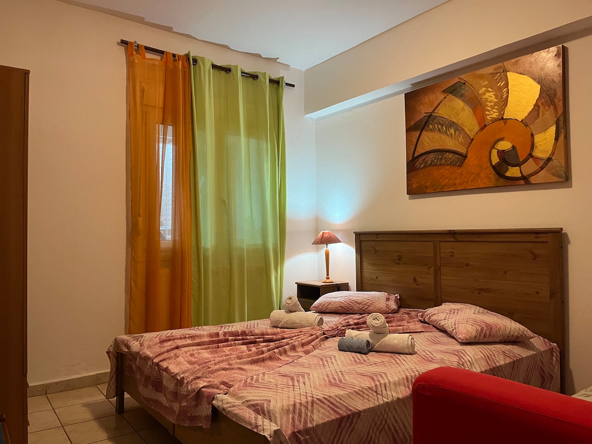 A spacious sleeping area is highlighted by patterned bedding and soft towels. The room features a wooden headboard and a bedside table with a lamp. Natural light enters through partially draped colorful curtains in green and orange, with a decorative wall art piece enhancing the space.
