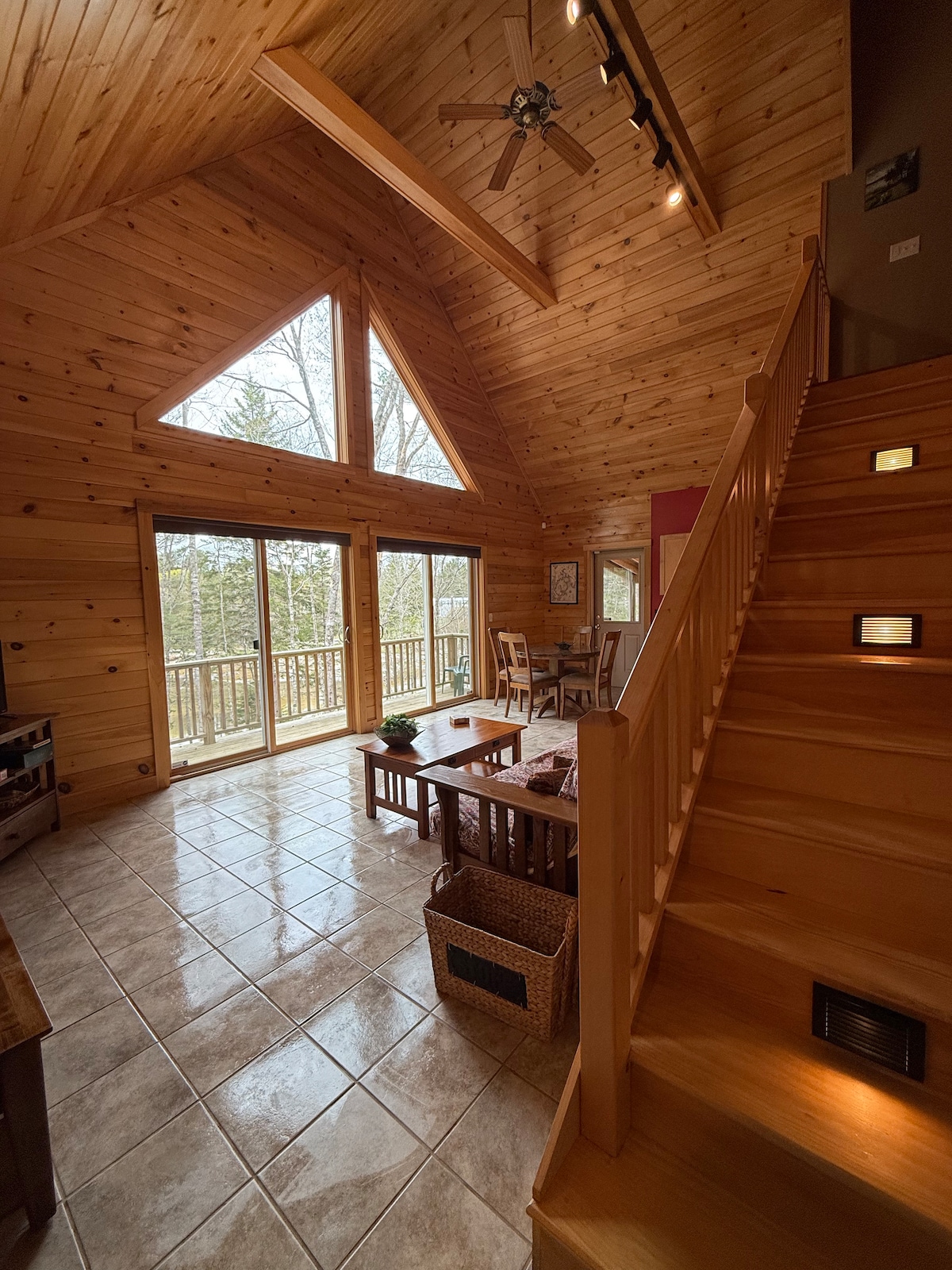 The spacious living area features high wooden ceilings and an abundance of natural light streaming through large triangular windows. Two sets of sliding glass doors lead to a deck, while a staircase with wooden railings invites movement to the loft area above. Comfortable furnishings are arranged for relaxation.