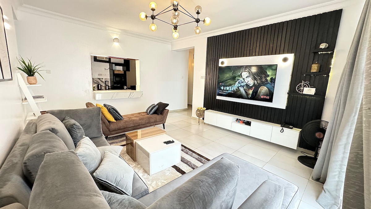 A modern living area features a comfortable gray sectional sofa and a stylish wooden coffee table. A large flat-screen television is mounted on a textured wall. Soft lighting from a contemporary chandelier illuminates the space, while a small plant adds a touch of greenery.
