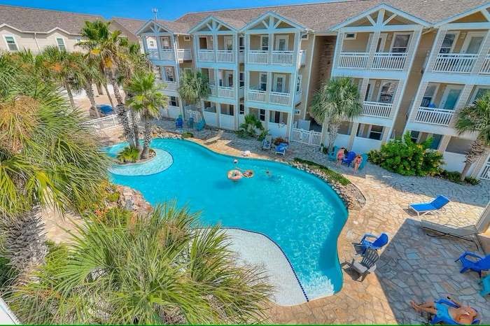 A resort-style pool is surrounded by palm trees and lounge chairs, providing a relaxing atmosphere. The pool's shape is inviting, with areas for swimming and shallow lounging. Two levels of balconies from the adjacent townhome overlook the scenic setting, enhancing the communal vacation experience.