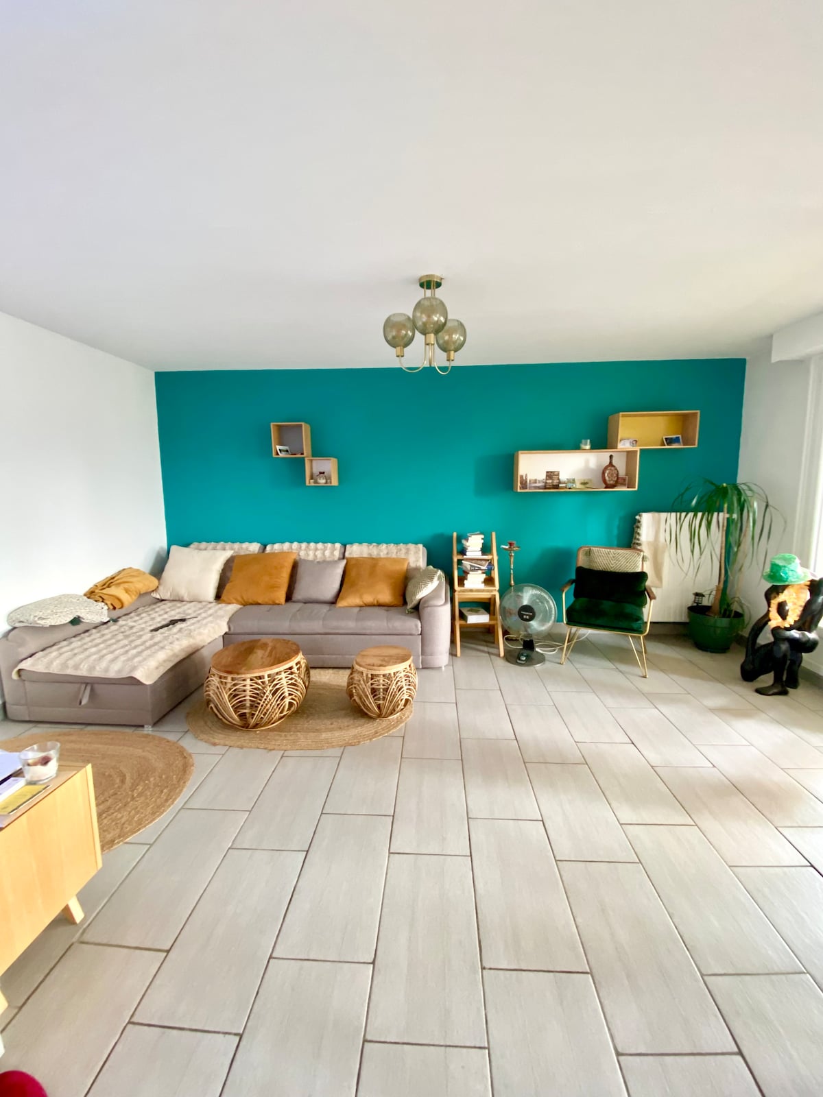 A bright living area is depicted, featuring a gray sofa adorned with cushions and a throw blanket. Decorative shelves are mounted on a vibrant teal wall, while two round wicker stools sit on the tiled floor, creating a cozy and inviting space.