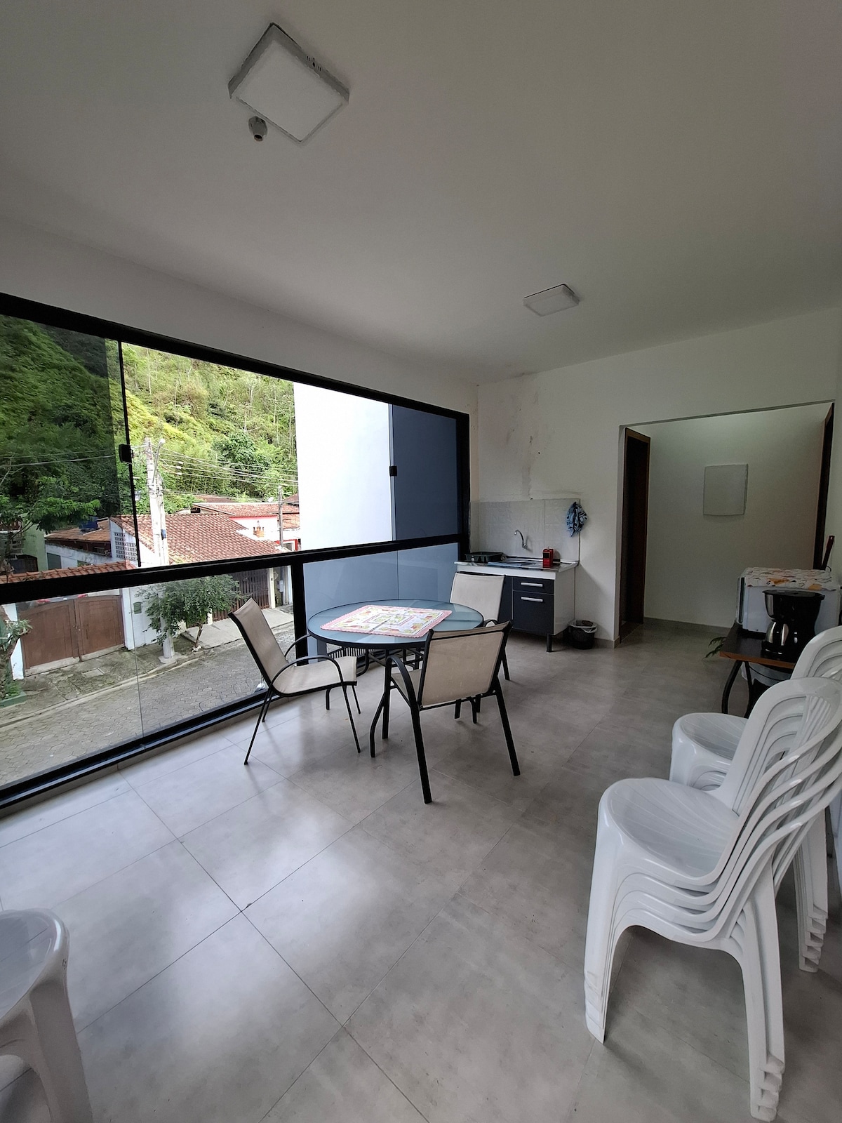 A bright, spacious area showcases a dining table surrounded by four chairs. A kitchenette is visible, equipped with essential appliances. Expansive glass walls provide ample natural light and an unobstructed view of the surrounding greenery, creating a serene atmosphere.