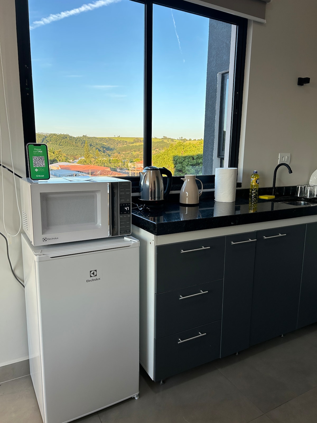A modern kitchen area features a compact refrigerator, a microwave, and an electric kettle, all positioned beside a sleek countertop. Natural light floods in through a large window, offering an open view of the surrounding landscape.