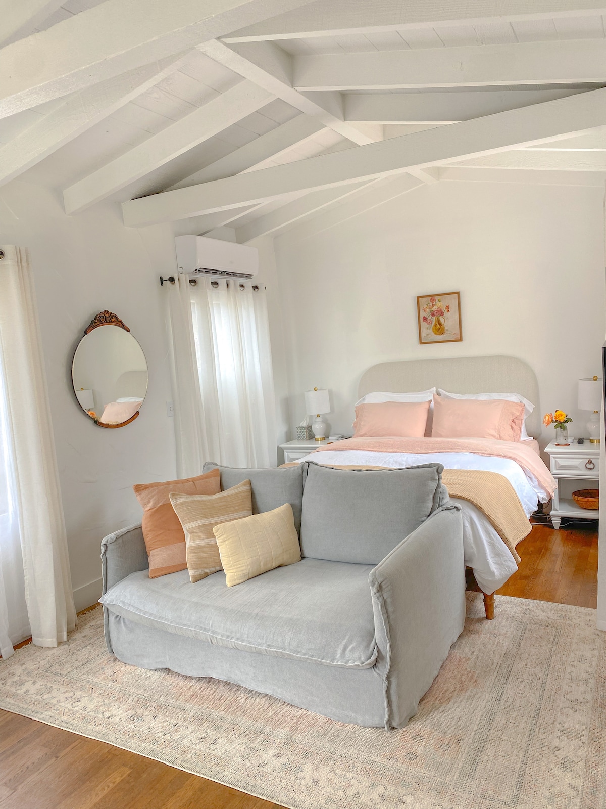 A cozy sleeping area features a spacious bed dressed in soft, natural fabric sheets. A light grey sofa is positioned in front of the bed, adorned with warm-toned throw pillows. Natural light filters through semi-sheer curtains, illuminating the room's vaulted wood beam ceilings.
