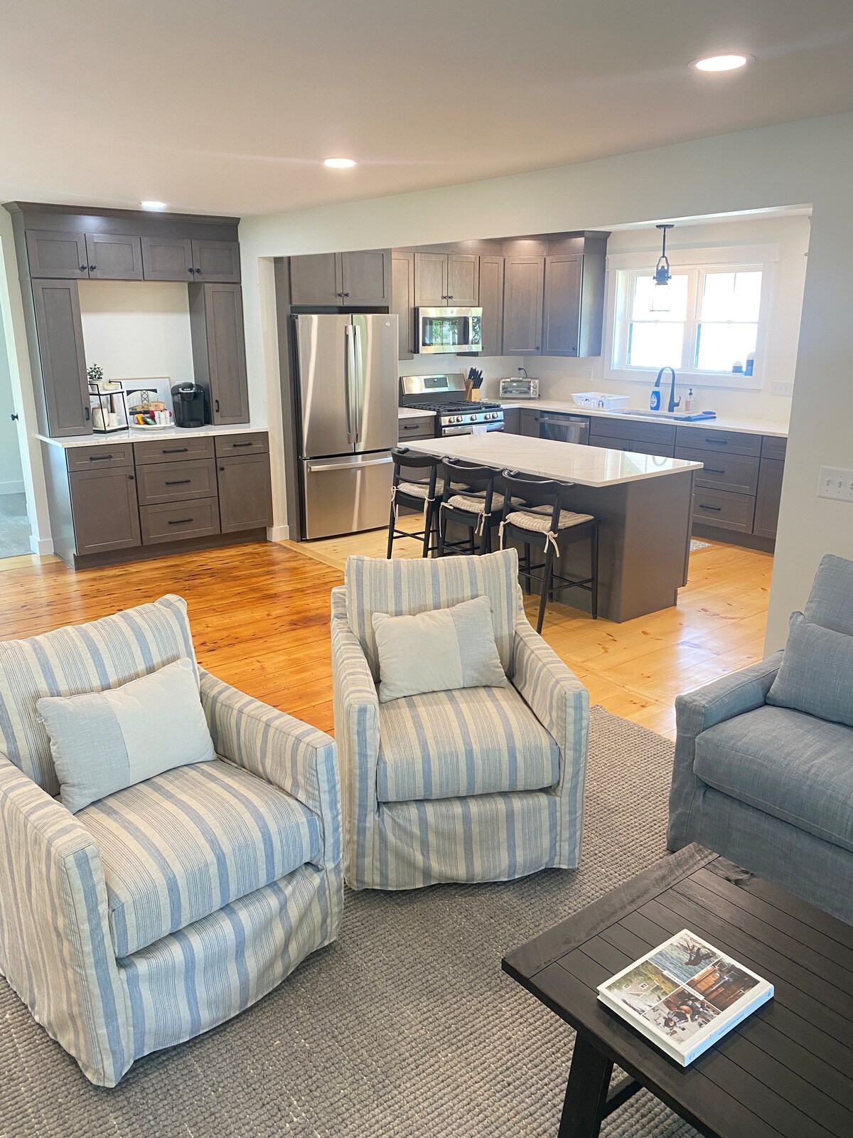 The fully renovated kitchen features modern appliances including a refrigerator and stove, with a central island providing additional prep space and seating. Cozy striped chairs offer a comfortable seating area, enhancing the open layout and inviting natural light.