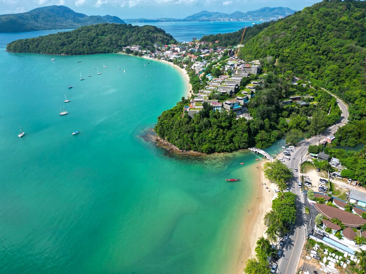 Aerial view of Ao Yon beach showcases a crescent-shaped sandy shoreline bordered by lush green hills. Various boats are anchored in serene turquoise waters, while a small community of colorful buildings is nestled along the coastline, highlighting the natural beauty of the area.