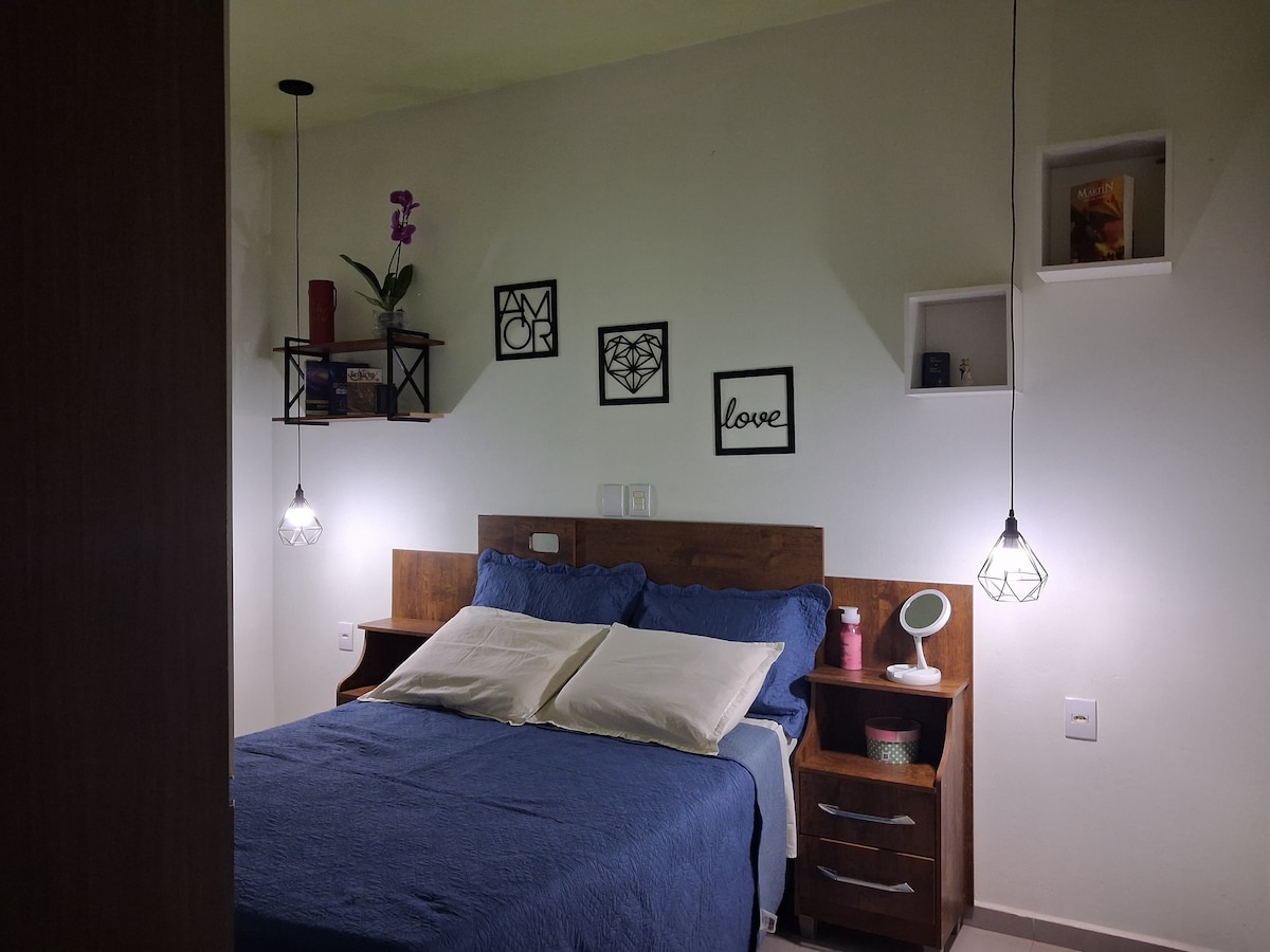 A sleeping area is presented with a spacious bed dressed in a blue coverlet, complemented by white pillows. Soft lighting is provided by pendant lamps on either side. Decorative shelves display a plant and framed artwork, enhancing the room's harmonious design.