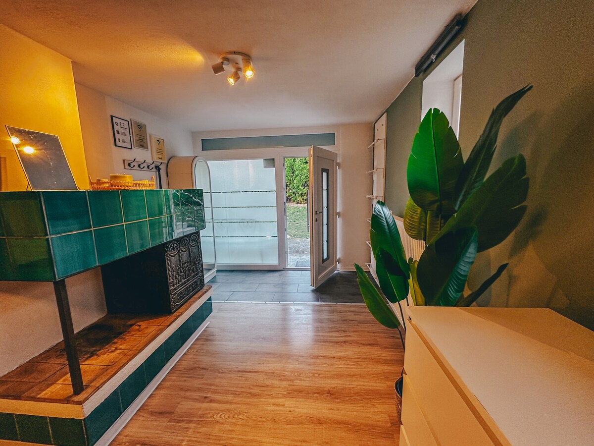 An inviting living space features a warm wooden floor, with a rich green tiled fireplace as a focal point. Natural light enters through double doors that open to an outdoor area, enhanced by potted greenery along the side wall.