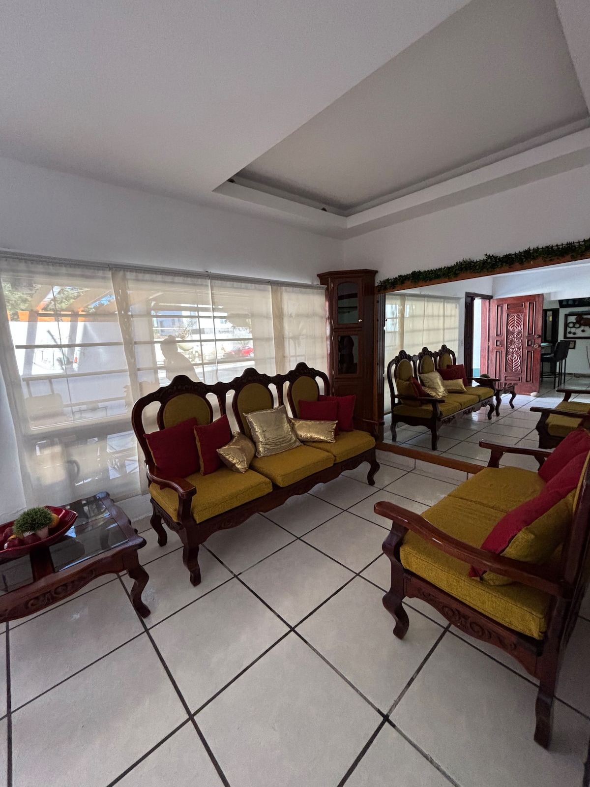 A bright living area features a set of wooden chairs with yellow upholstery and red cushions, arranged around a small table. Large windows allow natural light to fill the space, with sheer curtains softly filtering the view. A decorative mirror enhances the room's openness.