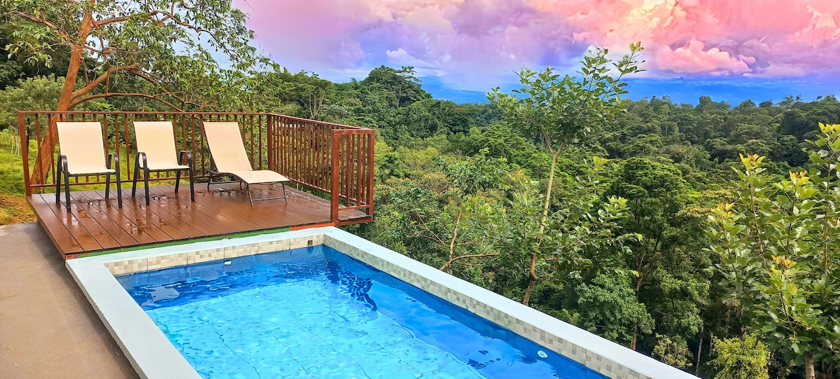 A secluded outdoor terrace features a refreshing swimming pool surrounded by lush greenery. Two lounge chairs are arranged on a wooden deck, with panoramic views of the forest and mountains in the distance. The soft colors of a sunset cast a serene glow across the scene.