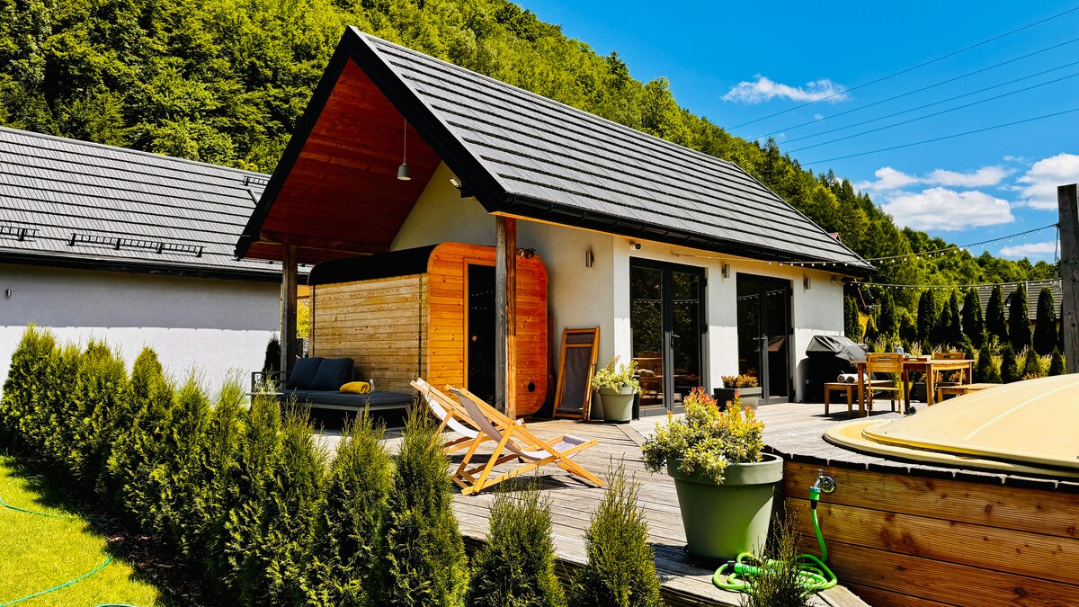 A modern retreat is surrounded by lush greenery and features a wooden sauna adjacent to the main structure. A spacious deck is furnished with outdoor seating and a table, inviting relaxation. The garden is complemented by neatly trimmed hedges and colorful potted plants.