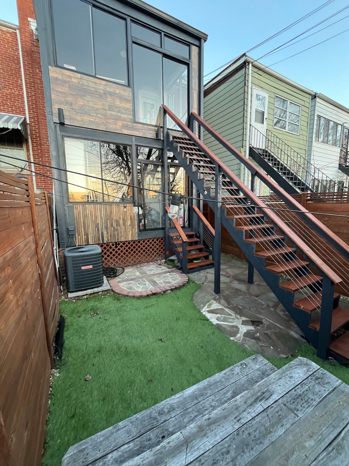 A small urban garden is depicted, featuring a wooden picnic table and an outdoor grill. A paved pathway leads to a stairway, providing access to the upper floor of the property. The garden is enclosed by vertical fencing and has a grassy area.