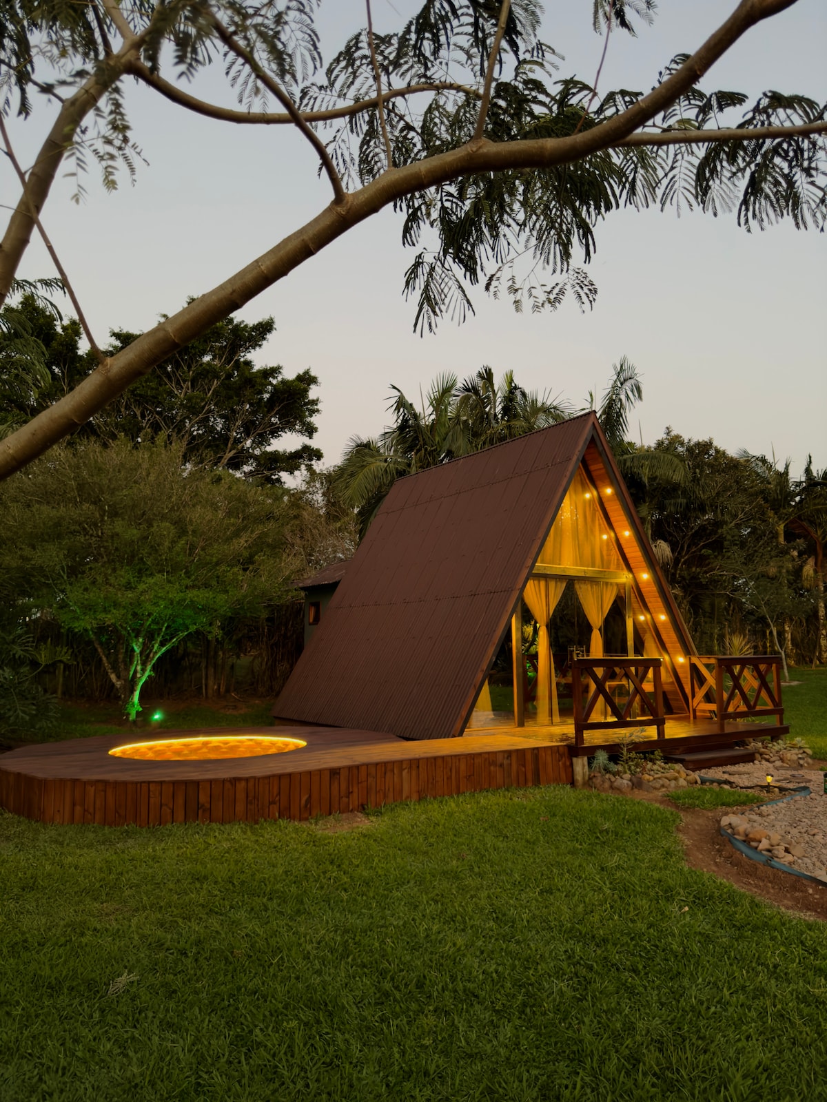 An A-frame cabin is set in a lush green area, showcasing soft exterior lighting around the deck and a circular hot tub on the side. The warm glow emanating from the cabin's large windows suggests a welcoming interior, surrounded by tall trees and plants.
