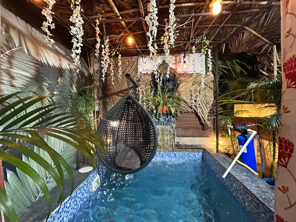 A hanging chair is seen beside a small water feature within an enclosed area. Soft lighting enhances the serene ambiance. Indoor plants are positioned around the space, contributing to the natural atmosphere, while a blue-tiled pool reflects the surrounding decor.