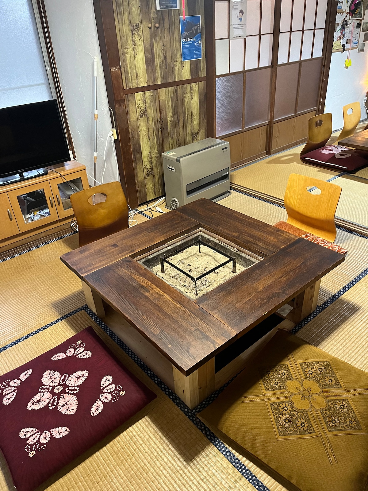 A traditional indoor area features a wooden irori table at the center, surrounded by several decorative floor cushions. A television and a heater are positioned along the wall, with tatami mats covering the floor. Shōji screens provide a glimpse of the room's Japanese design.