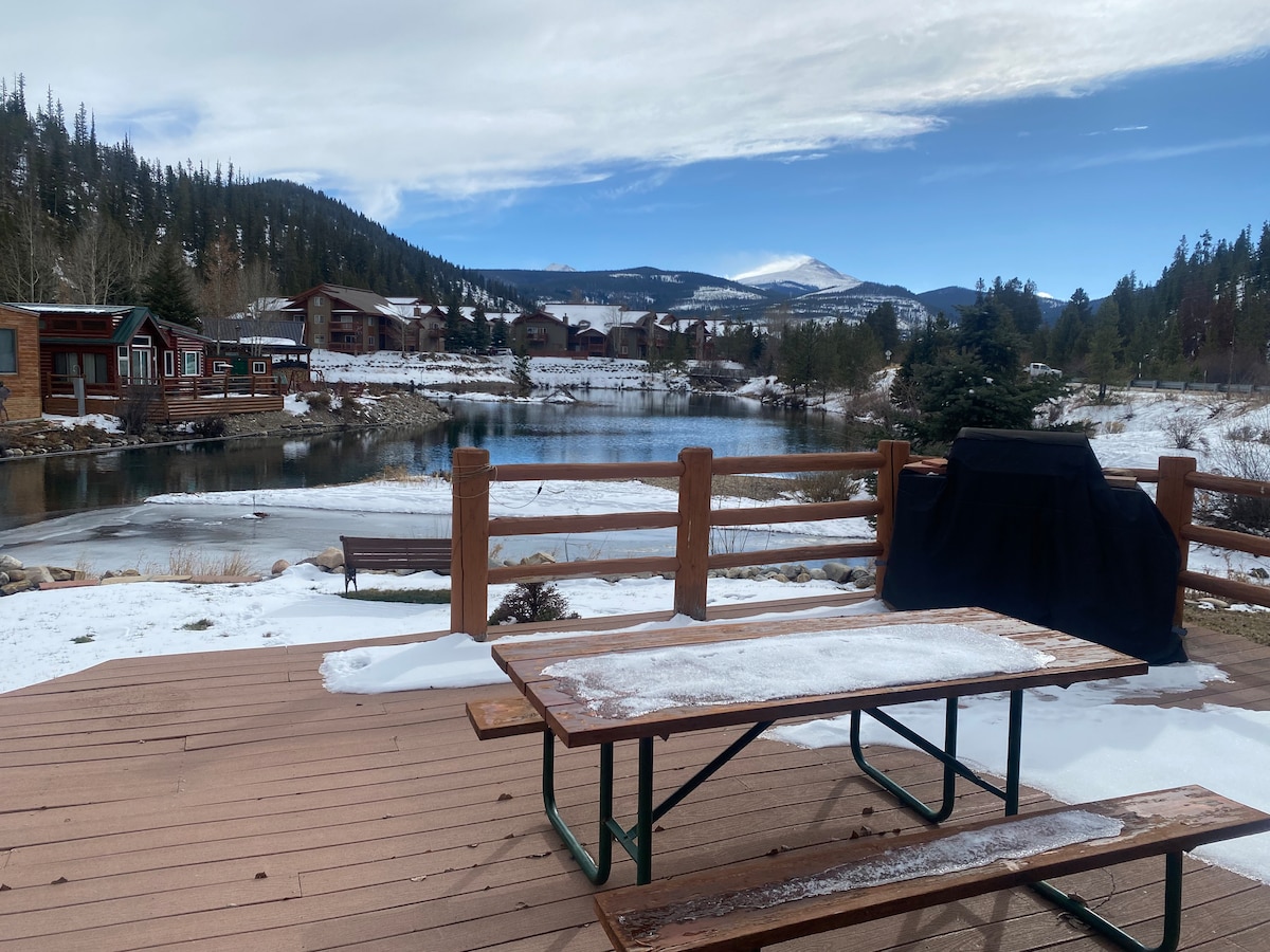 A private patio is set with a picnic table overlooking a serene lake surrounded by snow-dusted mountains. A grill is positioned nearby, and a snow-covered deck adds to the tranquil winter scene.