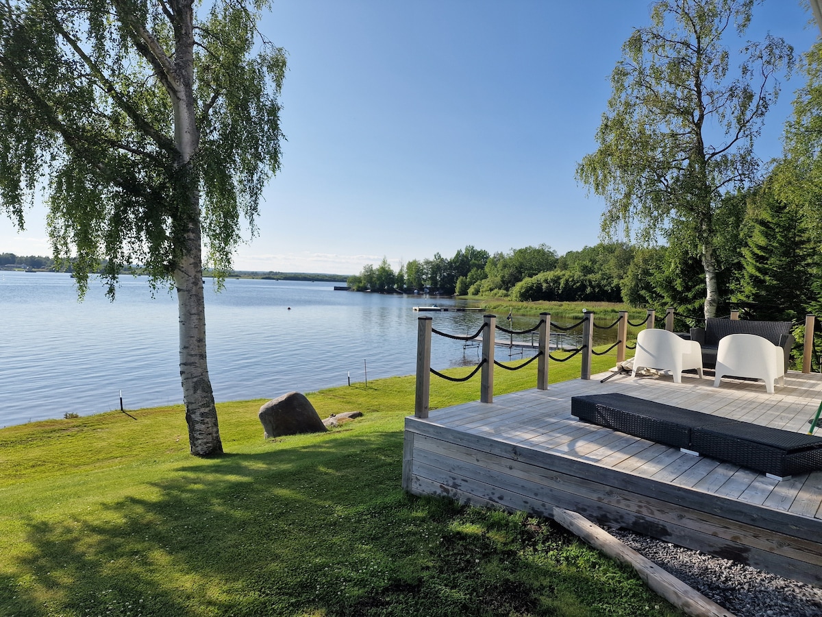 A serene waterfront view is presented, featuring a smooth lake surface reflecting the sky. A wooden deck is visible, with modern white chairs positioned nearby. Lush green grass and trees frame the scene, enhancing the sense of tranquility.