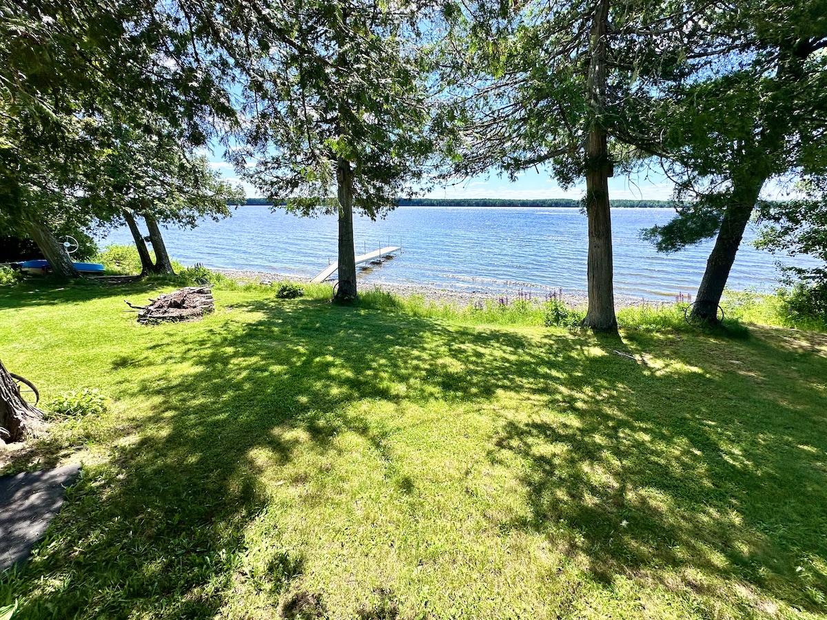 A lush green lawn gently slopes down to Cross Lake, framed by tall trees that provide dappled shade. The serene water stretches into the distance, with a wooden dock extending into the lake, inviting relaxation. The clear sky above hints at a pleasant day.