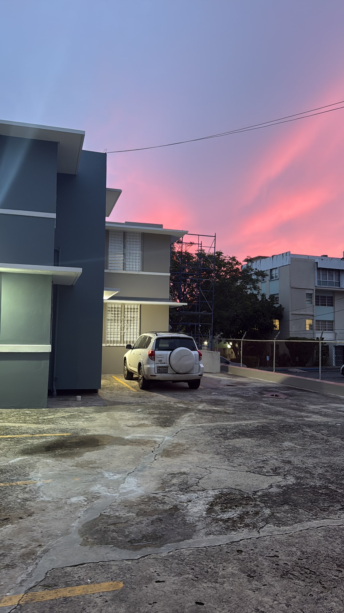 A modern building exterior is shown, characterized by clean lines and a muted color palette. A parked white vehicle is visible on the concrete driveway, with a backdrop of a colorful sunset sky casting soft hues over the scene.