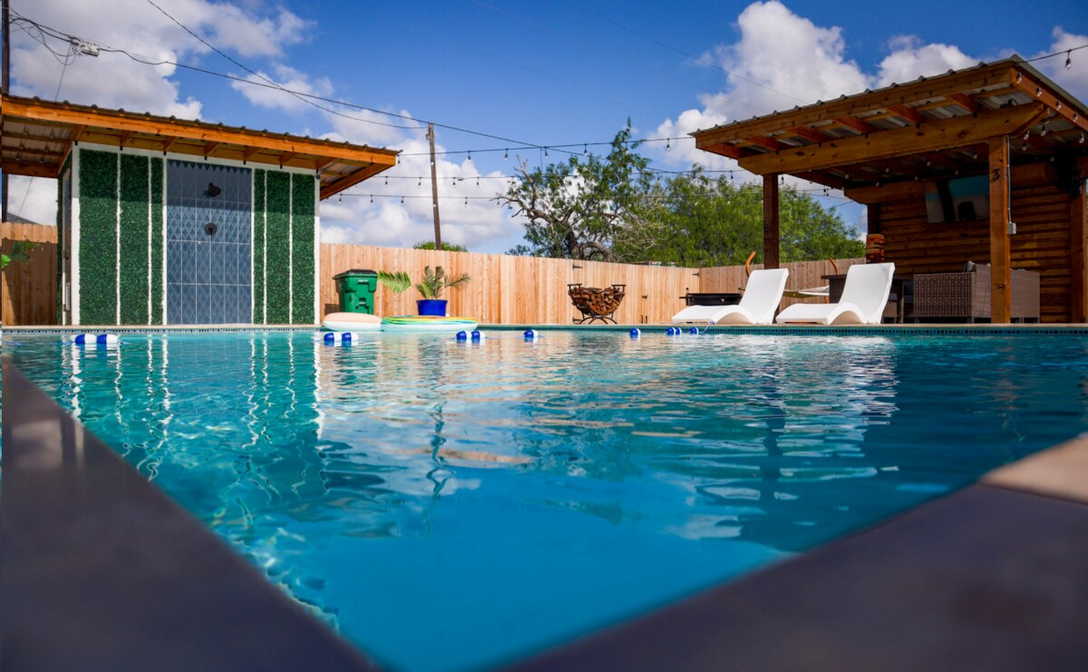 A large outdoor swimming pool glimmers under a clear blue sky, surrounded by a wooden deck. Two lounge chairs are positioned beside the pool, and a wooden structure offers shaded seating. A dressing area with a large window and a privacy fence is visible in the background.