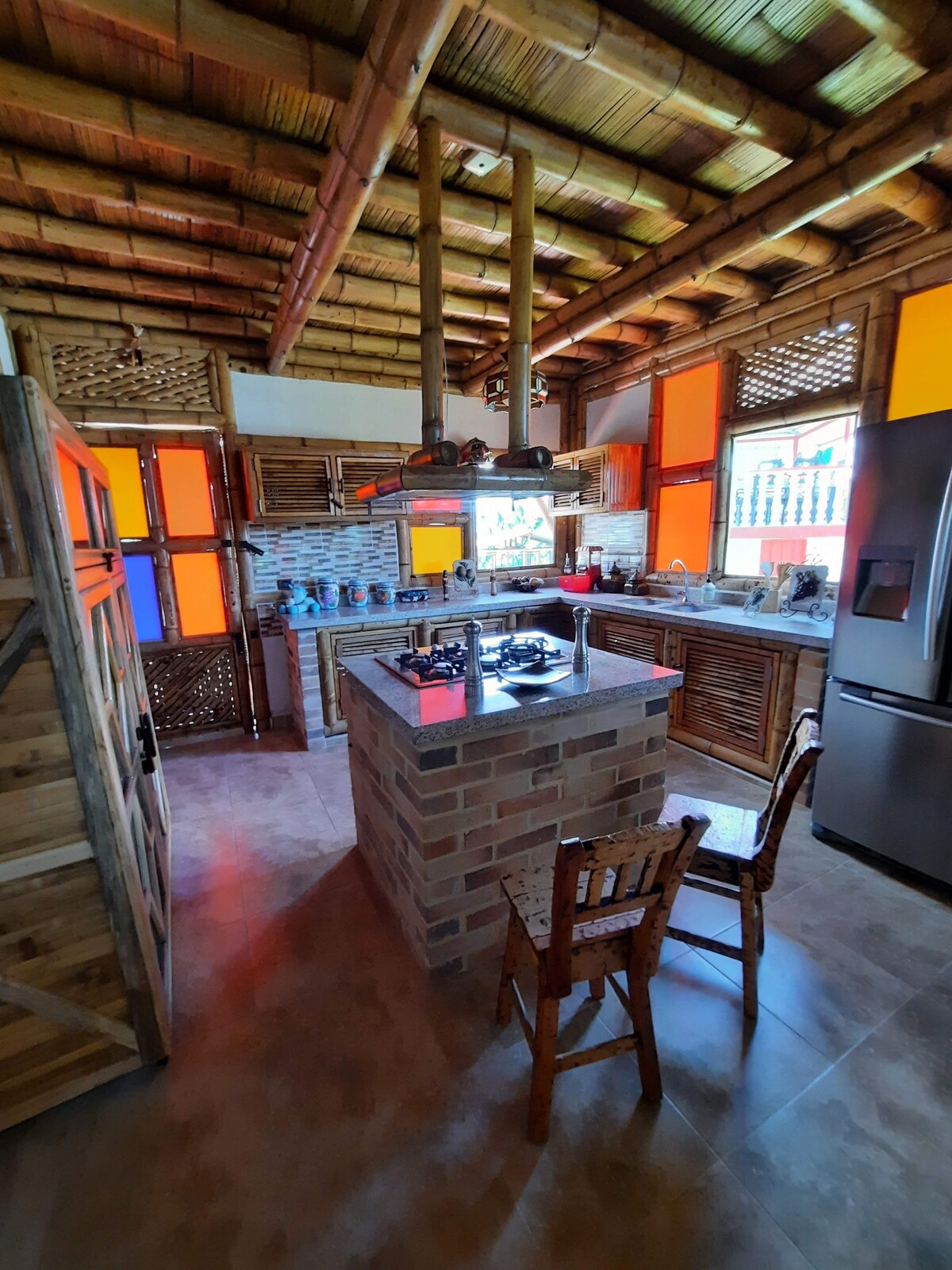 A rustic kitchen features bamboo ceilings and vibrant orange and blue windows, providing natural light. A central island with a stovetop is surrounded by wooden cabinetry. A small wooden chair is positioned nearby, while appliances seamlessly integrate into the design.