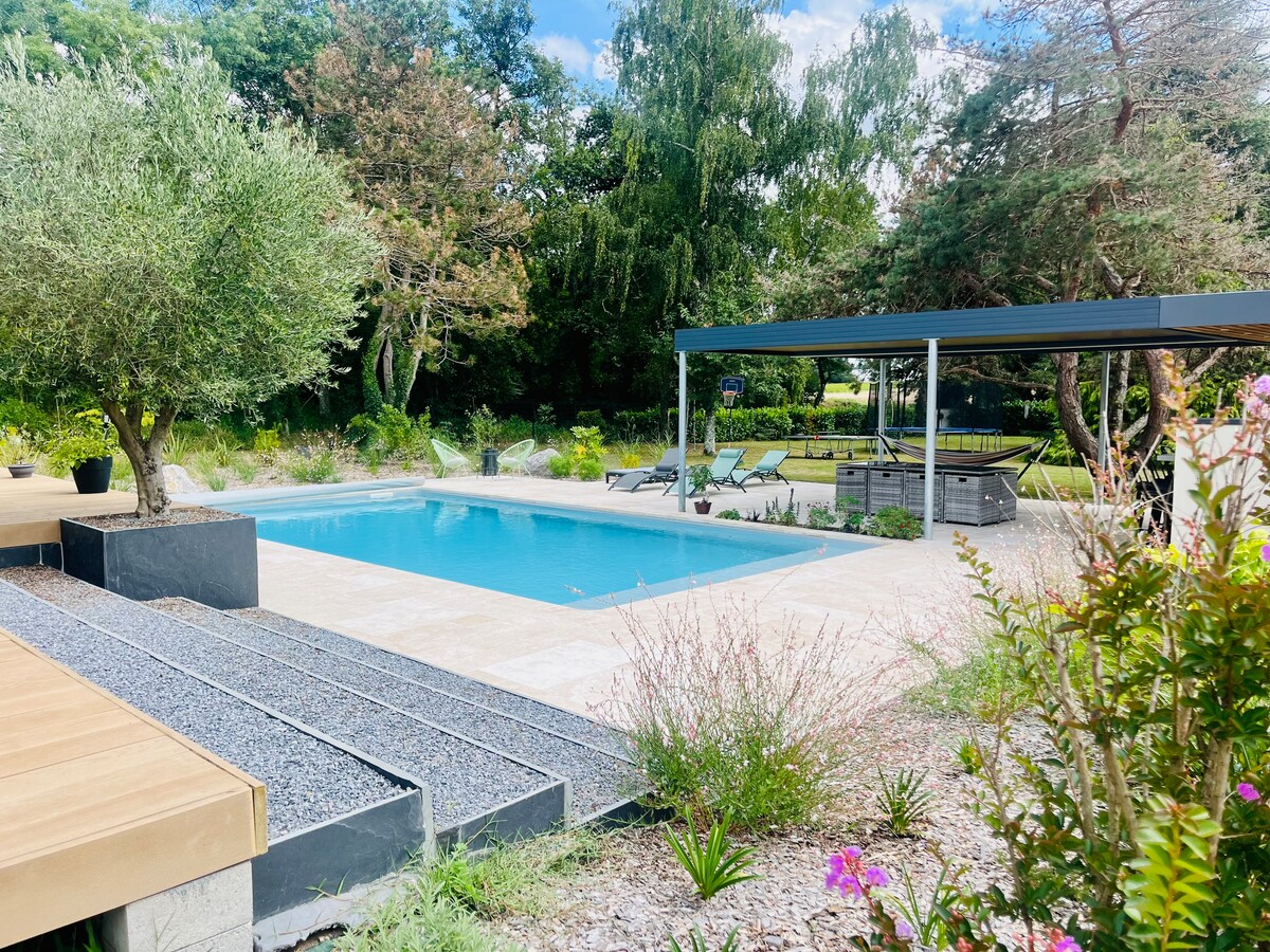 A private pool reflects the clear sky, surrounded by lush greenery and blooming flowers. Comfortable lounge chairs are positioned nearby under a shaded pergola, offering a serene space for relaxation. The pool area is enhanced by a smooth stone deck and tastefully arranged landscaping.