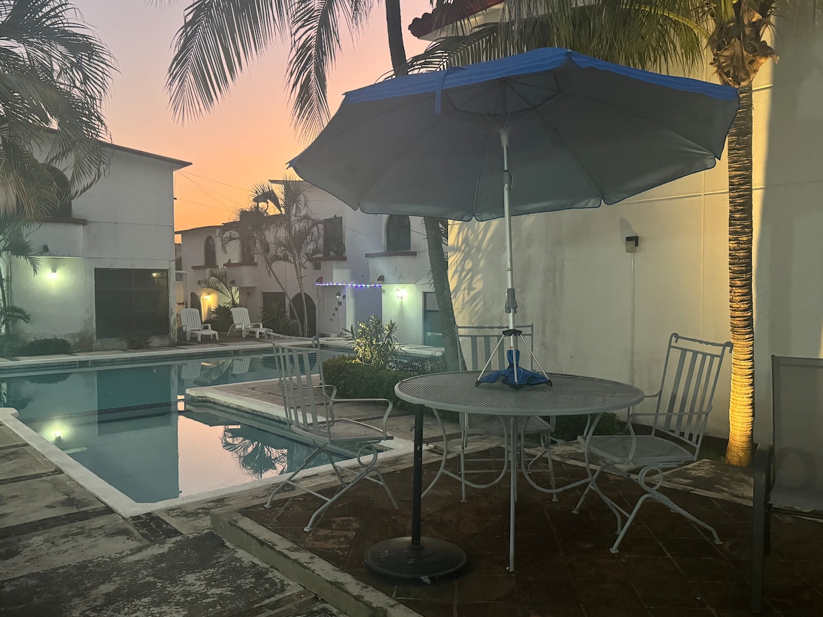 A shaded outdoor dining area features a round table surrounded by four chairs, positioned near a calm pool reflecting the soft evening light. Lush palm trees and well-maintained landscaping enhance the peaceful setting.