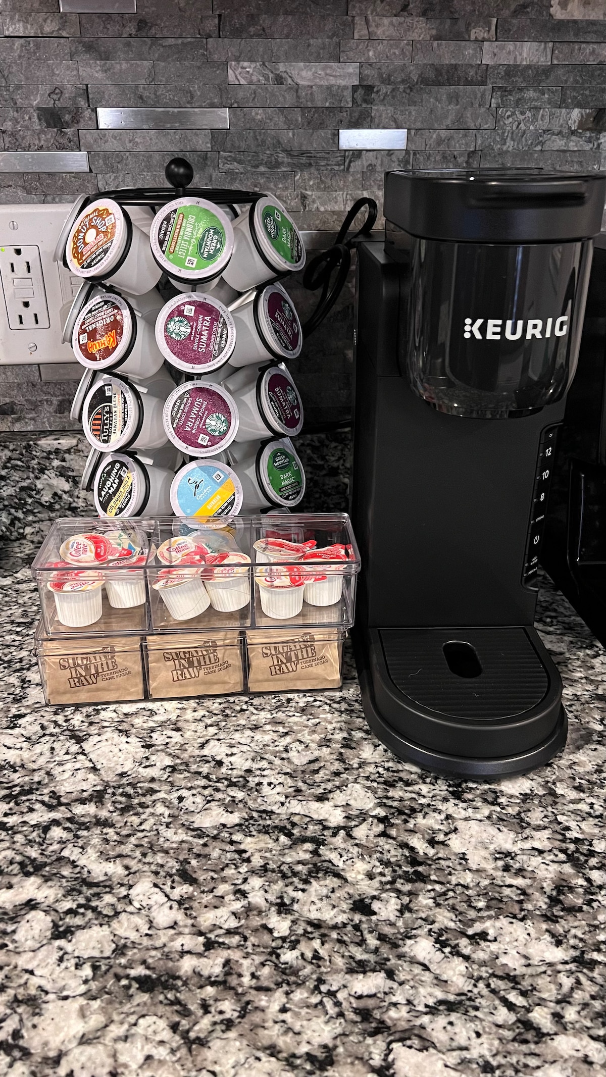 A coffee station is presented with a Keurig machine positioned next to a transparent storage container filled with coffee pods. A carousel holds multiple flavored coffee capsules within easy reach, all set against a backdrop of textured granite countertops.