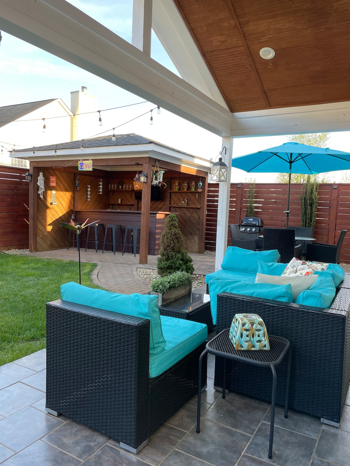 A relaxing backyard space features comfortable seating with bright, cushioned chairs. A wooden bar structure stands in the background, equipped with lighting and decorative elements. An umbrella provides shade over the seating area, surrounded by neatly trimmed grass and decorative landscaping.