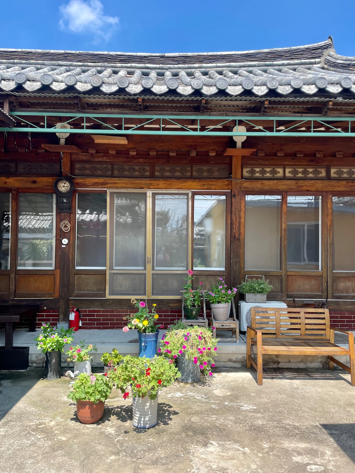 A traditional home with wooden beams and a tiled roof is featured. Potted plants with vibrant flowers decorate the entrance area. A wooden bench sits on the right side of the image, providing a welcoming spot to relax. The overall setting reflects a serene outdoor space.