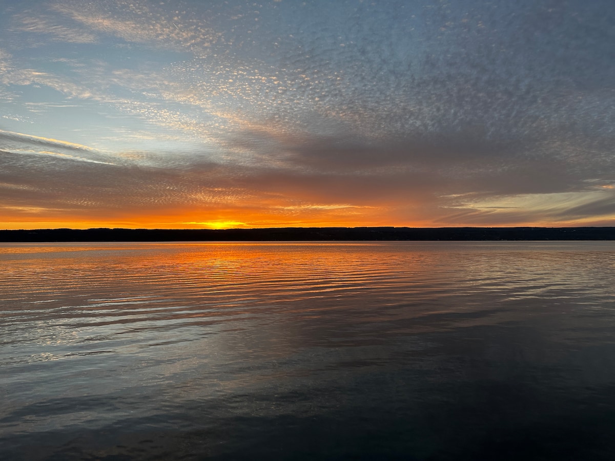 A serene sunset over Seneca Lake is captured, reflecting warm hues of orange and yellow across the calm water. The horizon is lined with distant hills under a softly clouded sky, creating a tranquil atmosphere that invites contemplation.