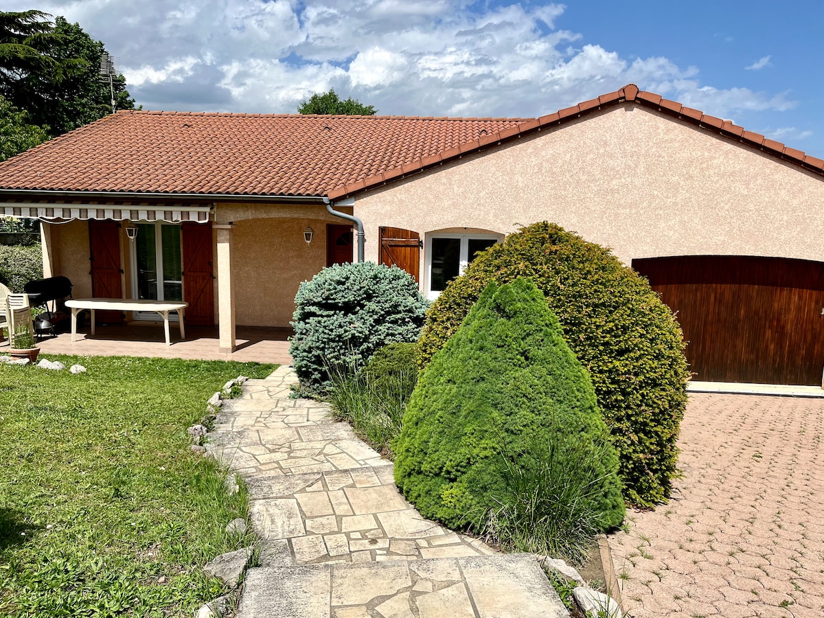 A single-story house features a warm exterior with a terracotta roof. A stone pathway leads to the entrance, bordered by well-maintained greenery, including shrubs and small trees. A covered porch with a table and chairs is visible, along with a garage on the side.