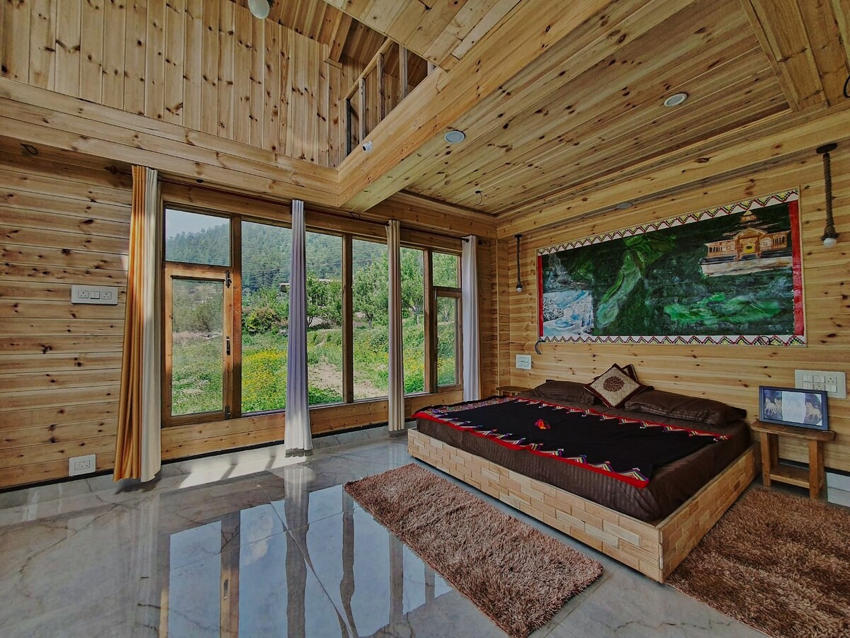 A spacious sleeping area features wooden paneling and large windows that provide views of the surrounding nature. A low platform bed is centered on a marble floor, accompanied by a colorful wall art piece. Soft rugs accentuate the room's cozy atmosphere.