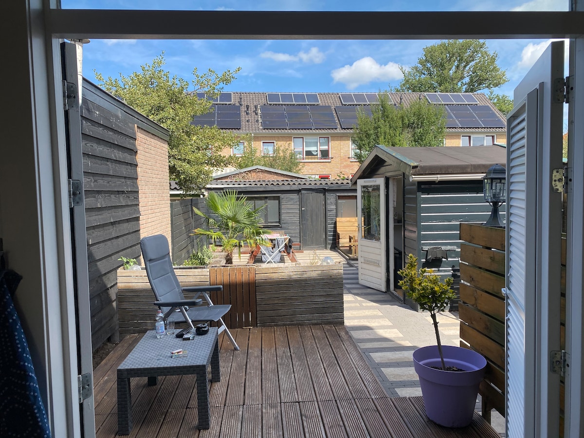 The outdoor space showcases a wooden deck with a seating area, surrounded by greenery and potted plants. A relaxed lounge chair is positioned beside a small table. In the background, solar panels are visible on rooftops, contributing to a serene environment.