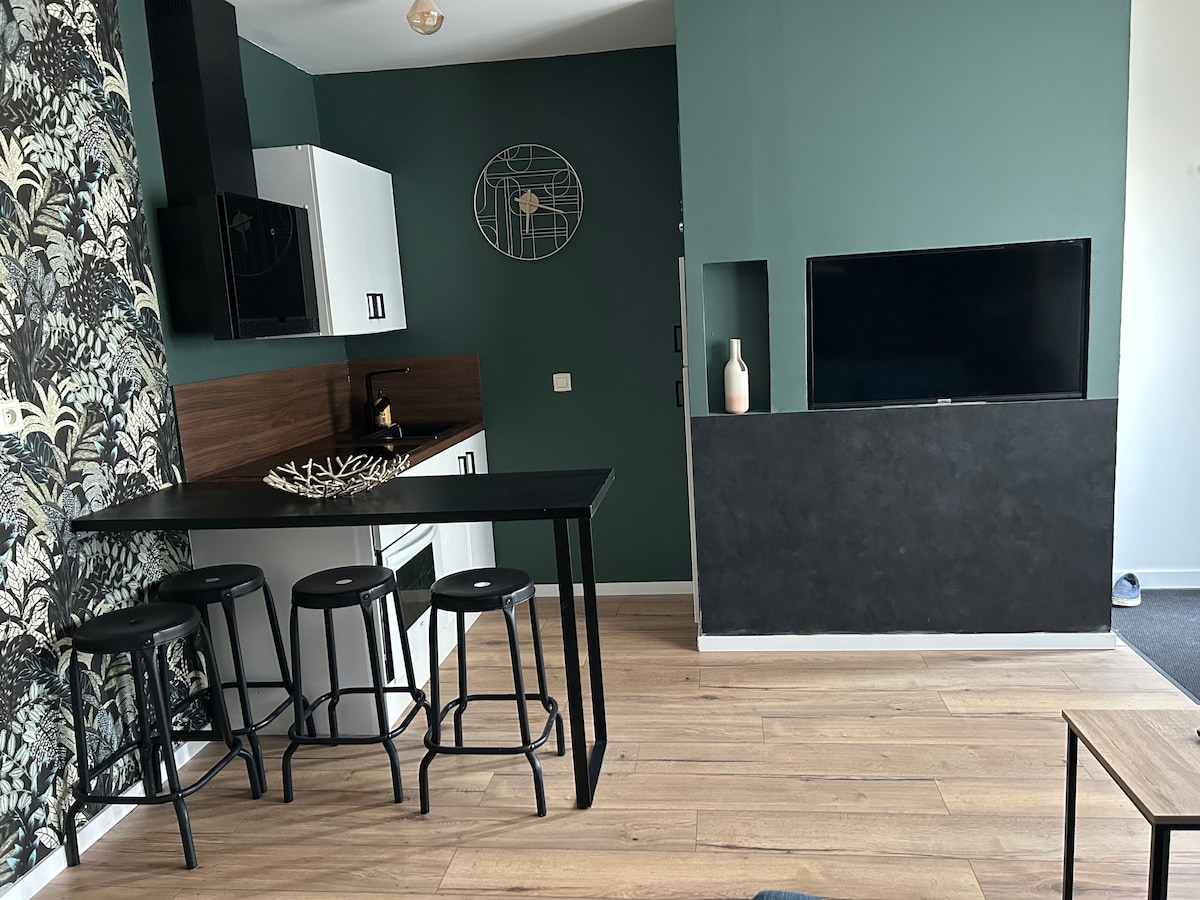 An open living area features a kitchen with white cabinetry and dark wood accents. A black bar table with three stools is positioned near the kitchen. A dark accent wall houses a television, providing a modern contrast to the green walls and patterned wallpaper.